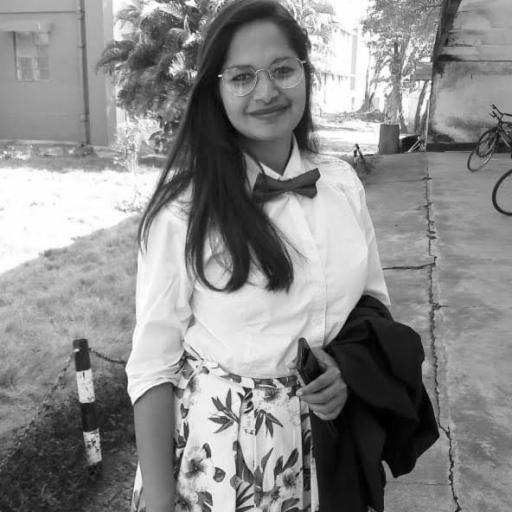 A brown girl in a white short and a printed skirt outside standing in grass. She is carrying her black blazer over her left arm.